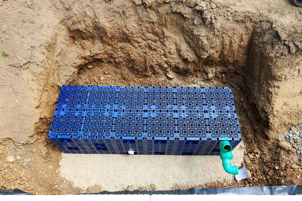 Blue geocellular crates bedded on gravel in an excavated trench, forming an attenuation soakaway, with the inlet pipe connected
